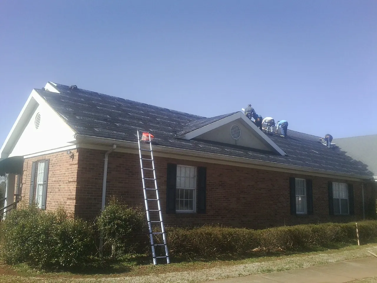 Crew tearing shingles off a brick ranch roof with a ladder set — Metal Roof Repair work by our crew in Georges Mills