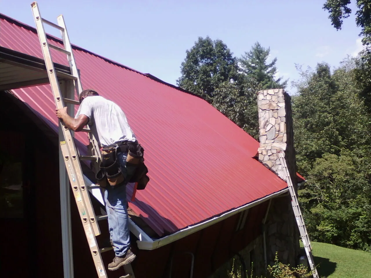 Roofer on a ladder beside a red metal roof and stone chimney — Soffit & Fascia Repair by Roofing Brothers in Georges Mills