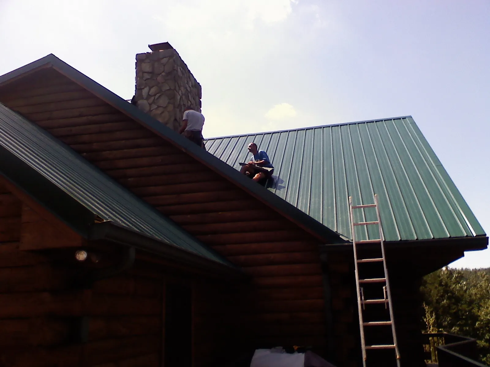 Roofers installing green metal panels beside a stone chimney on a log home in Georges Mills
